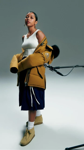 A fashionable woman stands in a studio wearing a tank top, navy skirt, tan boots, and a mustard-colored puffer jacket draped around her.