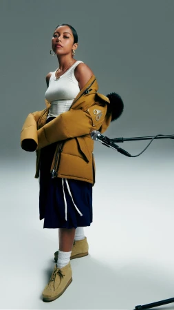 A fashionable woman stands in a studio wearing a tank top, navy skirt, tan boots, and a mustard-colored puffer jacket draped around her.