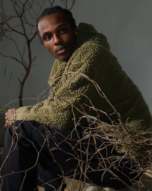 A person wearing a green sherpa jacket and dark pants is sitting amongst branches in a studio with soft lighting, looking at the camera.