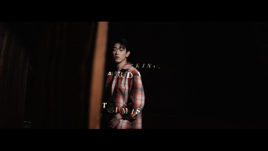 Young man stands in front of black backdrop, wearing a red and white plaid shirt, styled hair, and subtle rim lighting.