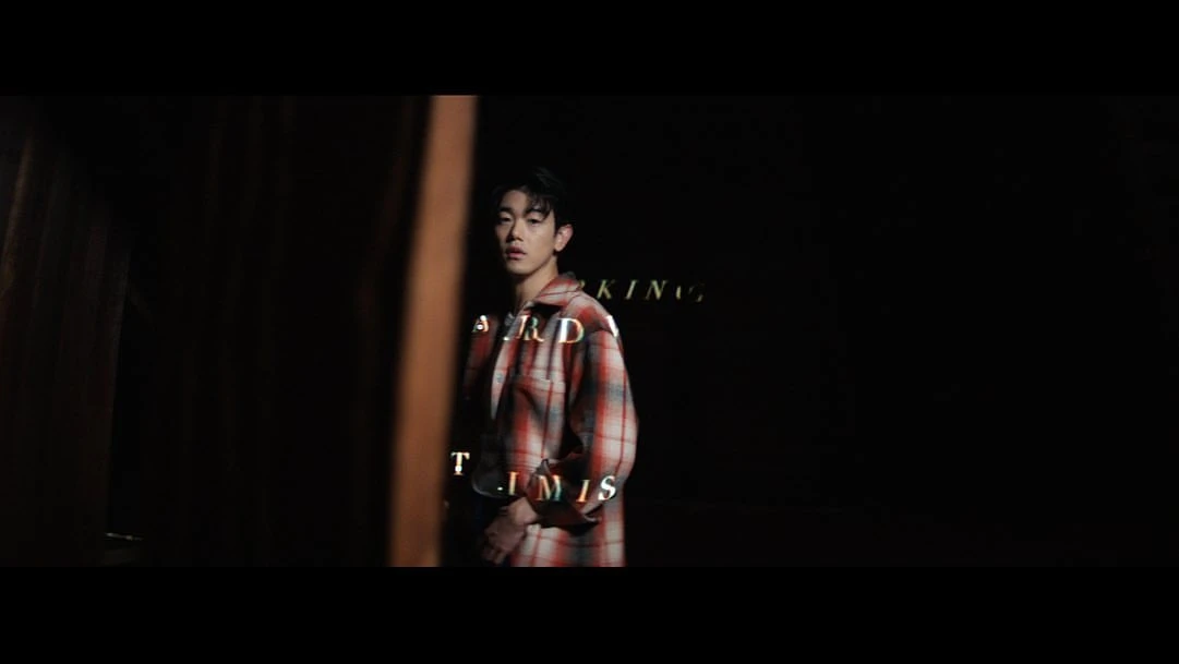 Young man stands in front of black backdrop, wearing a red and white plaid shirt, styled hair, and subtle rim lighting.