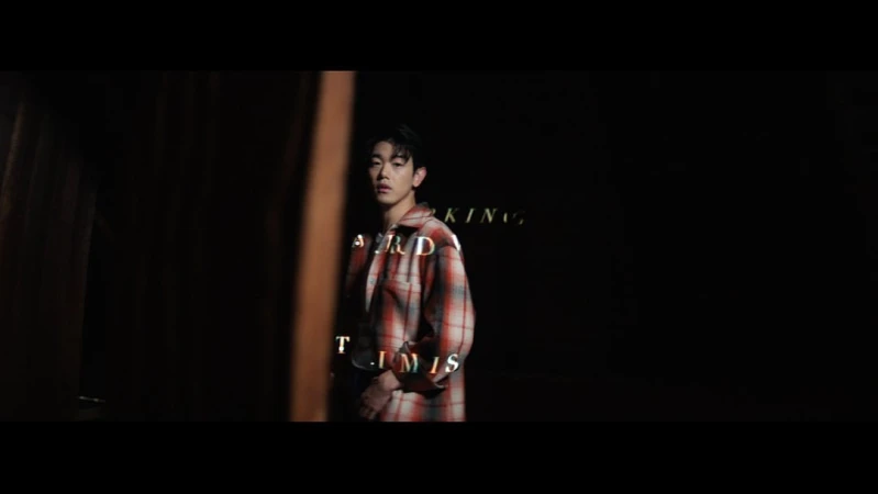 Young man stands in front of black backdrop, wearing a red and white plaid shirt, styled hair, and subtle rim lighting.