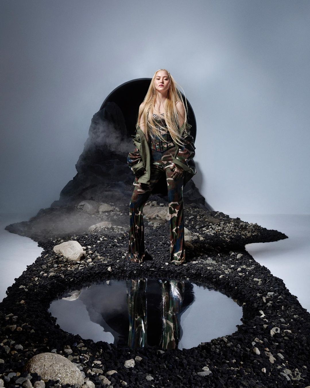 Woman in camouflage outfit posing confidently on a textured ground with a reflective pool, set against a smoky, cavernous backdrop.