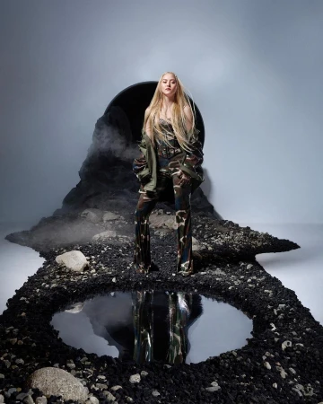 Woman in camouflage outfit posing confidently on a textured ground with a reflective pool, set against a smoky, cavernous backdrop.