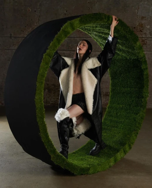 Fashion photo of a woman in a black and white coat posing inside a large green and black circular prop in a concrete setting.