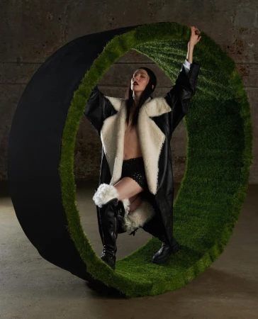 Fashion photo of a woman in a black and white coat posing inside a large green and black circular prop in a concrete setting.