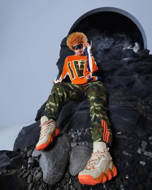 Ice Spice poses on a rocky surface wearing orange shirt, camo pants, orange sneakers, sunglasses with an orb/arch background.
