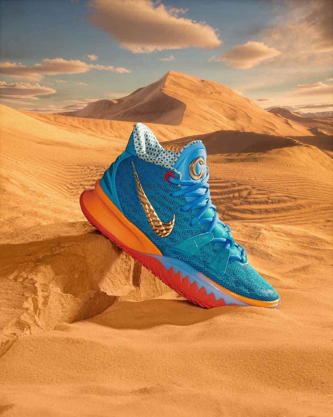 Turquoise and orange Nike Kyrie shoe resting on sand, with a desert landscape and dunes in the background under a cloudy sky.