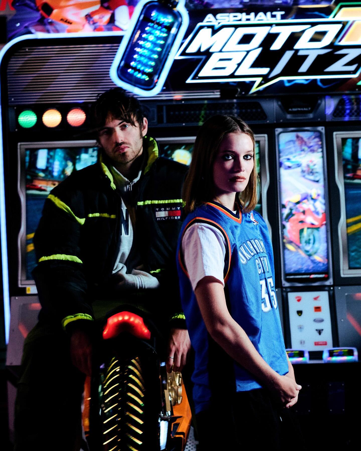 A man and a woman pose in front of an 'Asphalt Moto Blitz' arcade game, illuminated by colorful lights and a motorcycle visible.