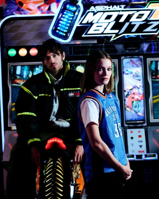 A man and a woman pose in front of an 'Asphalt Moto Blitz' arcade game, illuminated by colorful lights and a motorcycle visible.