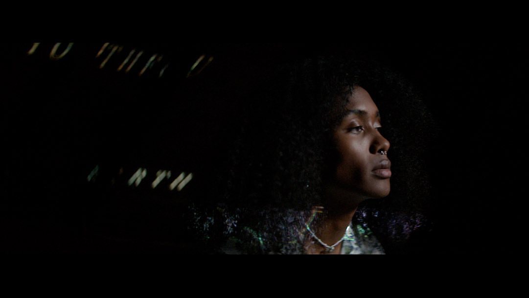 Close-up profile of an adult woman with curly hair and a nose ring, looking upward in dim lighting with obscured text in the background.