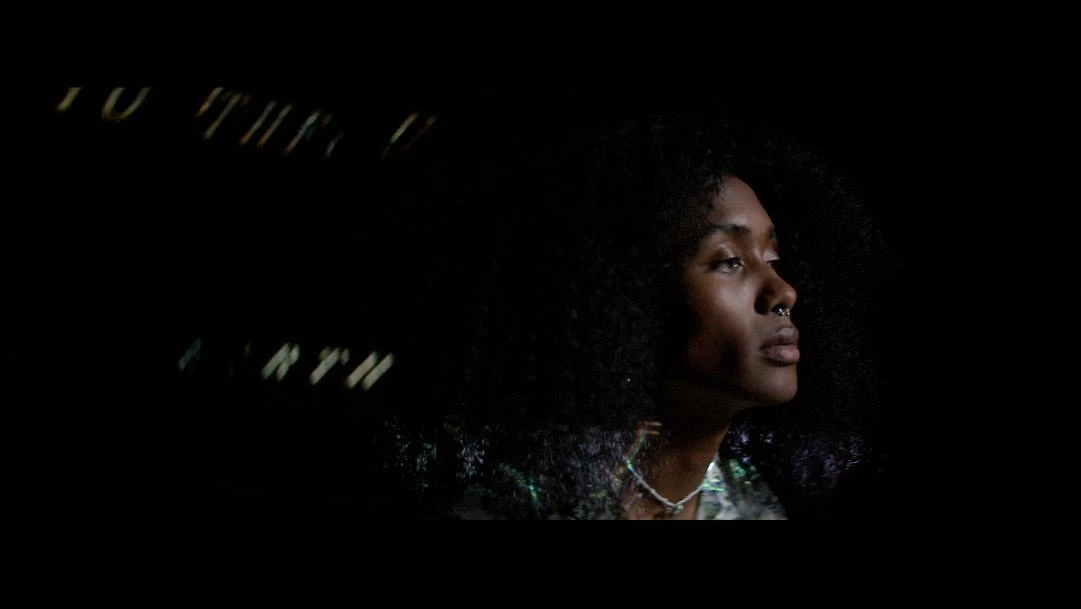 Close-up profile of an adult woman with curly hair and a nose ring, looking upward in dim lighting with obscured text in the background.