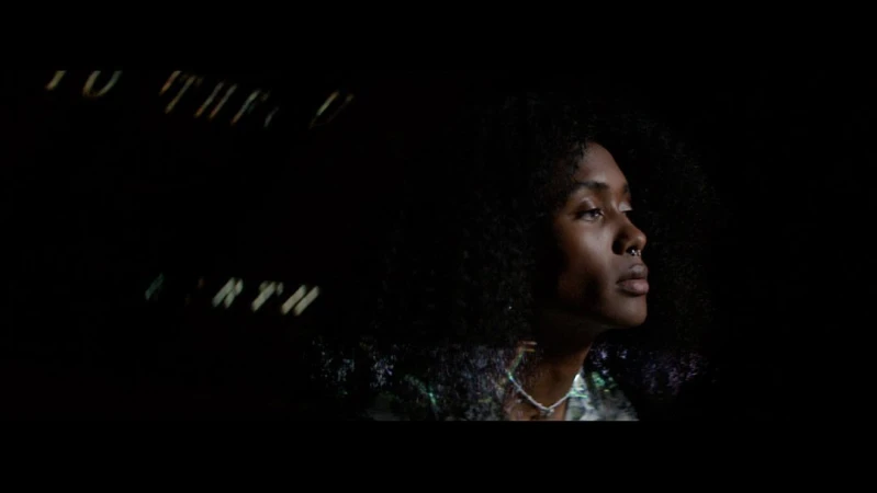 Close-up profile of an adult woman with curly hair and a nose ring, looking upward in dim lighting with obscured text in the background.