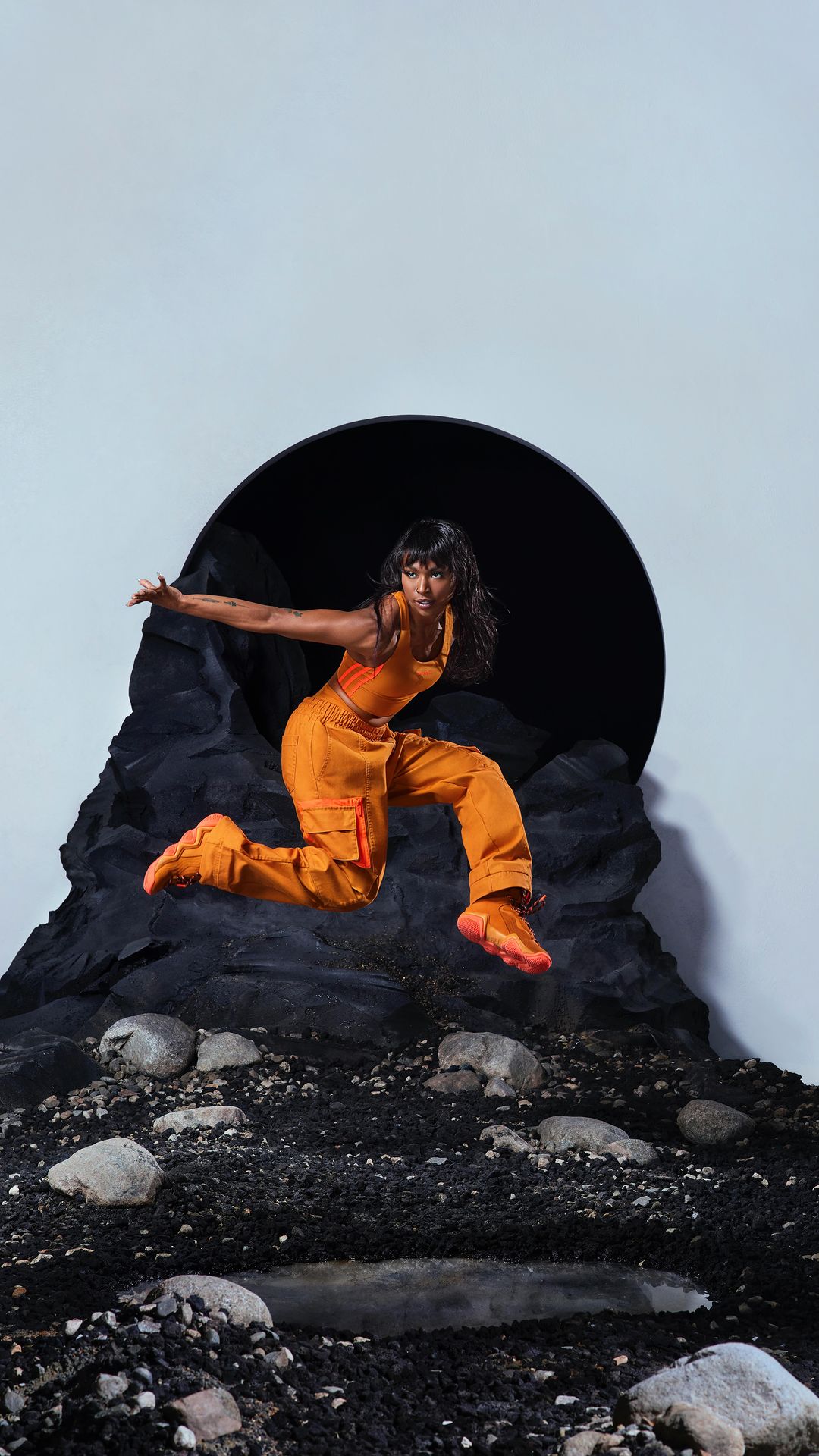 Athletic woman in orange sportswear powerfully leaps in a studio with gray rock formations and a dark portal backdrop, showcasing fitness and style.