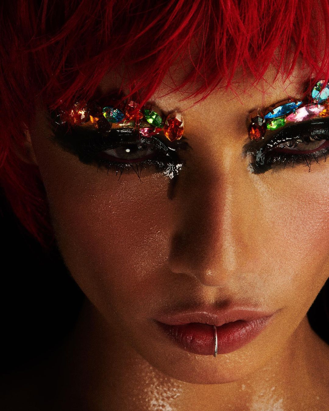 Close-up portrait of a woman with red hair, a lip ring, and dramatic makeup featuring black eyeliner and colorful jewels around the eyes.