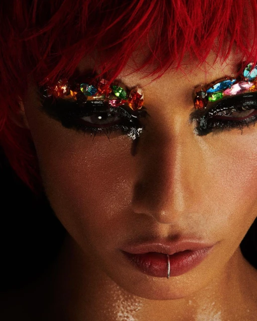 Close-up portrait of a woman with red hair, a lip ring, and dramatic makeup featuring black eyeliner and colorful jewels around the eyes.