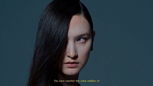 Portrait of a person with long, straight black hair partially covering their face, set against a muted blue-grey background.