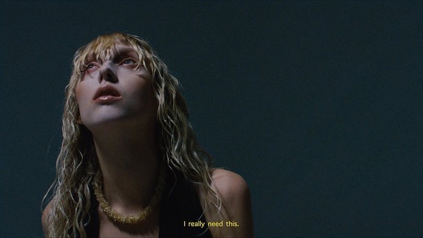 Close-up portrait of a woman with wet blonde hair wearing a beaded necklace, looking upwards against a dark background.