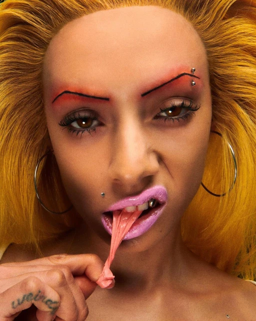 Close-up of a person with yellow hair and stylized makeup blowing bubblegum. The image focuses on the face, highlighting the makeup and expression.