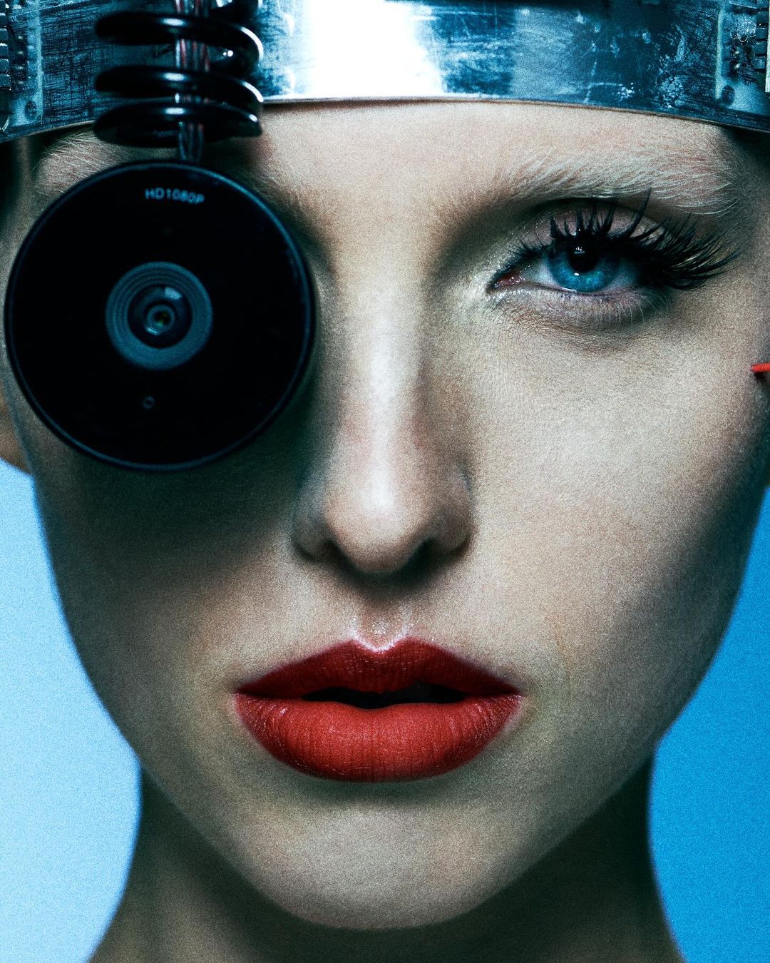Close-up of a woman with a metallic headpiece and a camera covering one eye, bright red lipstick, against a light blue backdrop.