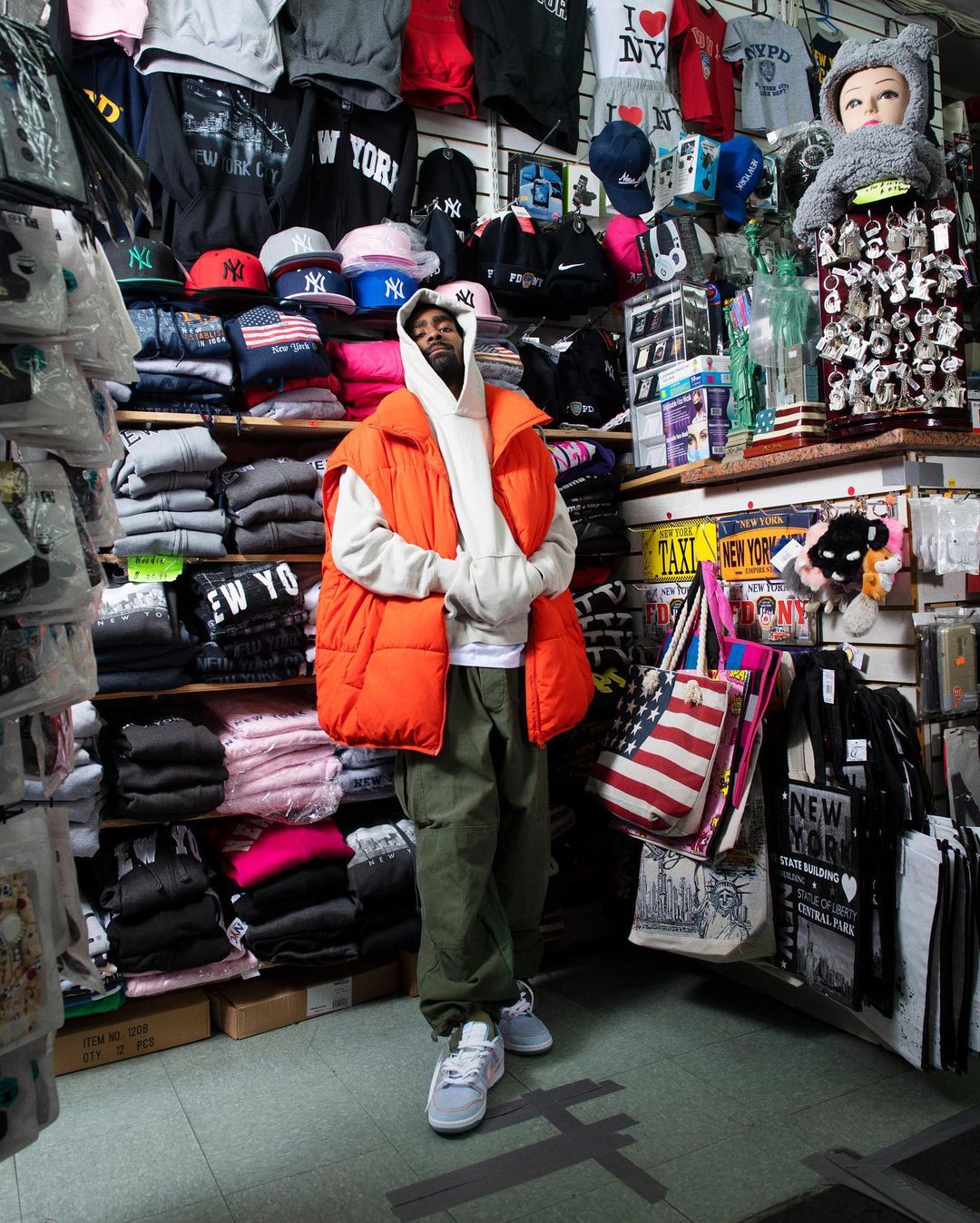 Man poses in orange puffer vest, hoodie, and sneakers in a store full of souvenirs and apparel, capturing a candid urban moment.