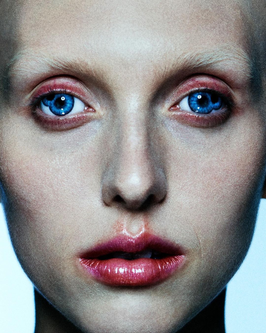 Close-up portrait of a fair-skinned adult female with bright blue eyes, red lipstick, and a pale complexion, featuring a beauty and makeup aesthetic.