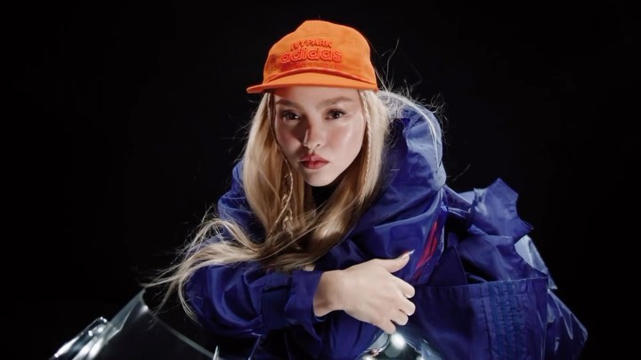 A person with fair skin and blonde hair wears a cap and jacket against a dark backdrop, shot in a fashion or editorial style.