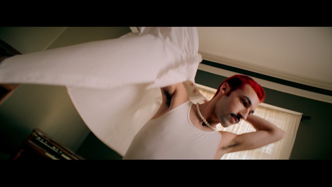 Low-angle view of a person with red hair holding a white sheet in a bedroom with tan walls, a pearl necklace, and a white tank top.