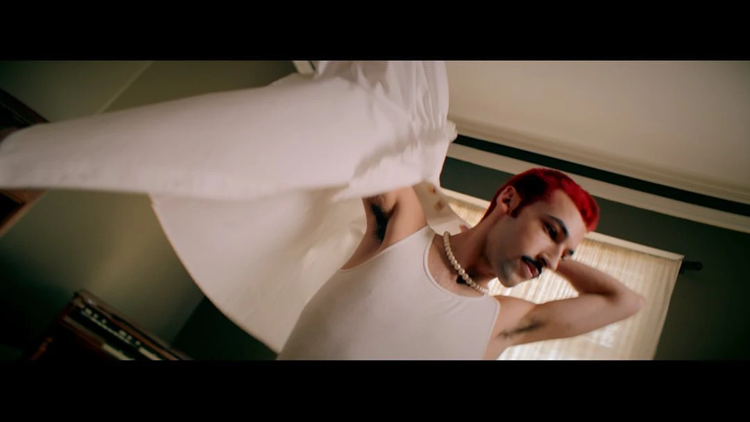 Low-angle view of a person with red hair holding a white sheet in a bedroom with tan walls, a pearl necklace, and a white tank top.