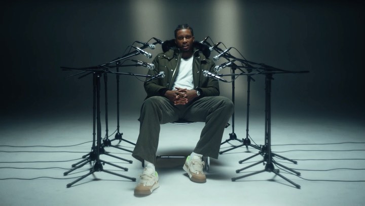Adult male sitting in a chair surrounded by microphones in a studio setting, wearing a jacket and sneakers, captured in a medium shot.
