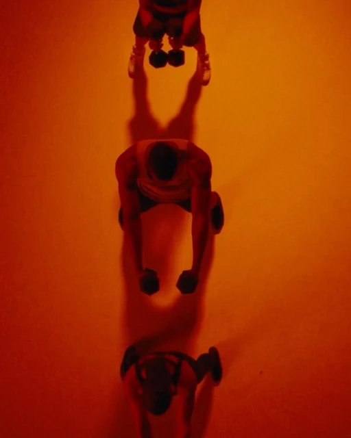 Overhead shot of three people in a line lifting dumbbells in a warm, orange-toned, evenly lit gym or studio setting.