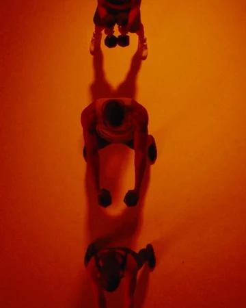 Overhead shot of three people in a line lifting dumbbells in a warm, orange-toned, evenly lit gym or studio setting.