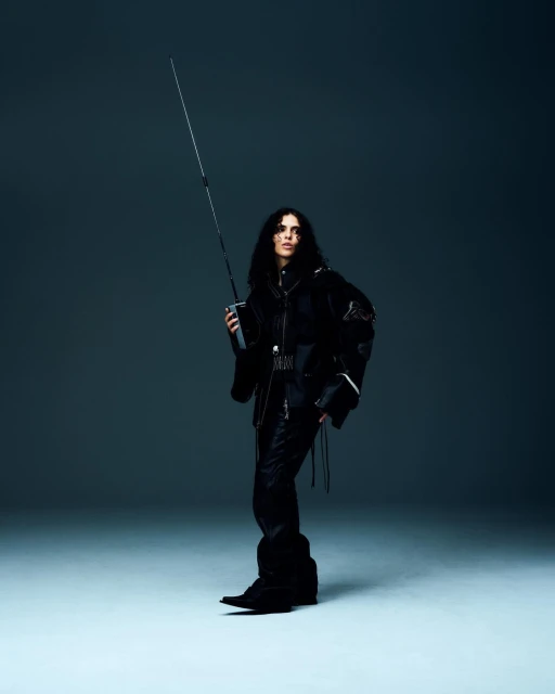 A female adult with long dark hair poses in a black outfit, holding a long black sword in a full studio shot against a grey backdrop.