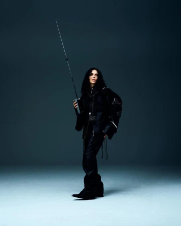 A female adult with long dark hair poses in a black outfit, holding a long black sword in a full studio shot against a grey backdrop.