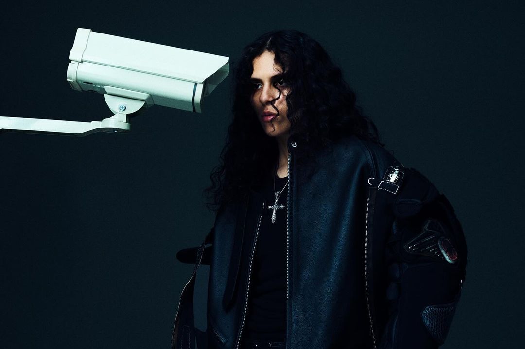 Portrait of a young adult man with curly hair in a leather jacket, posing next to a security camera against a dark background.