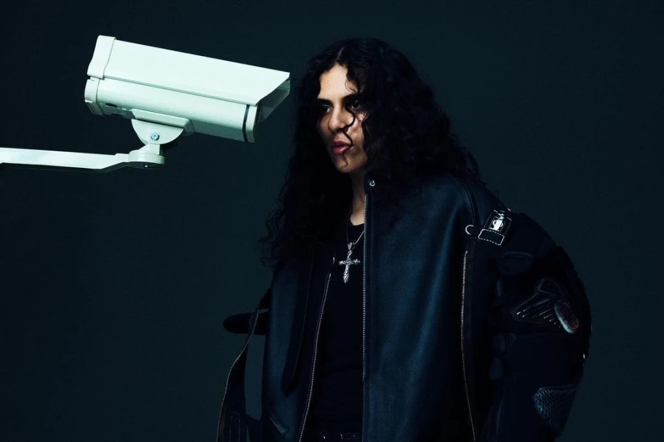 Portrait of a young adult man with curly hair in a leather jacket, posing next to a security camera against a dark background.