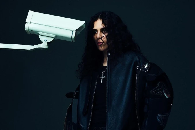 Portrait of a young adult man with curly hair in a leather jacket, posing next to a security camera against a dark background.