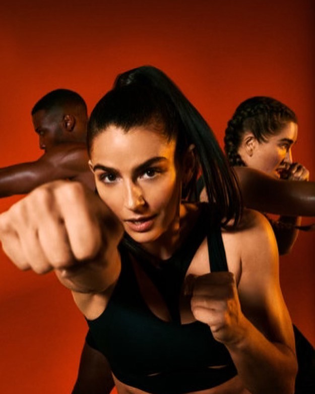 Three athletes in fitness attire pose in a boxing stance against a vibrant orange backdrop, highlighted by dynamic studio lighting.