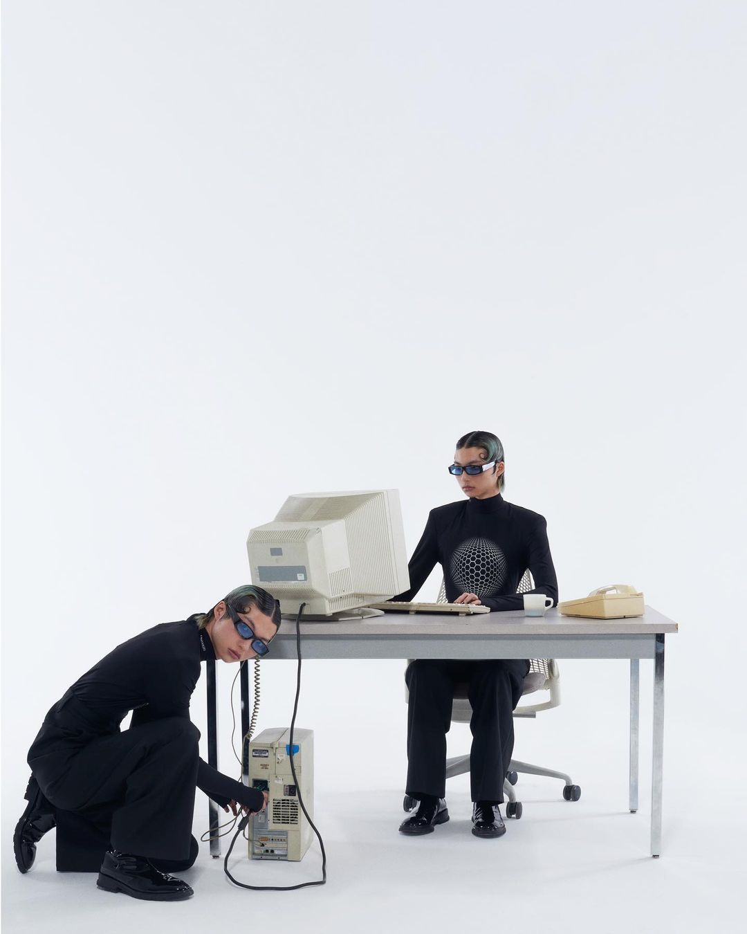 Two people in black outfits troubleshoot an old computer setup in a white studio, featuring vintage PC, monitor, desk, chair, and phone.