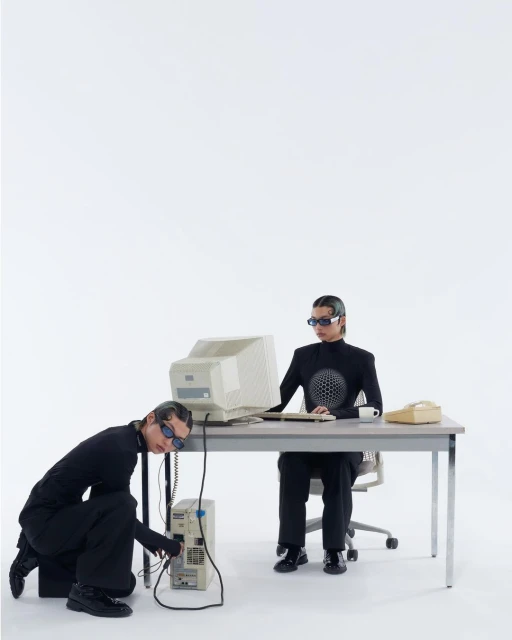 Two people in black outfits troubleshoot an old computer setup in a white studio, featuring vintage PC, monitor, desk, chair, and phone.