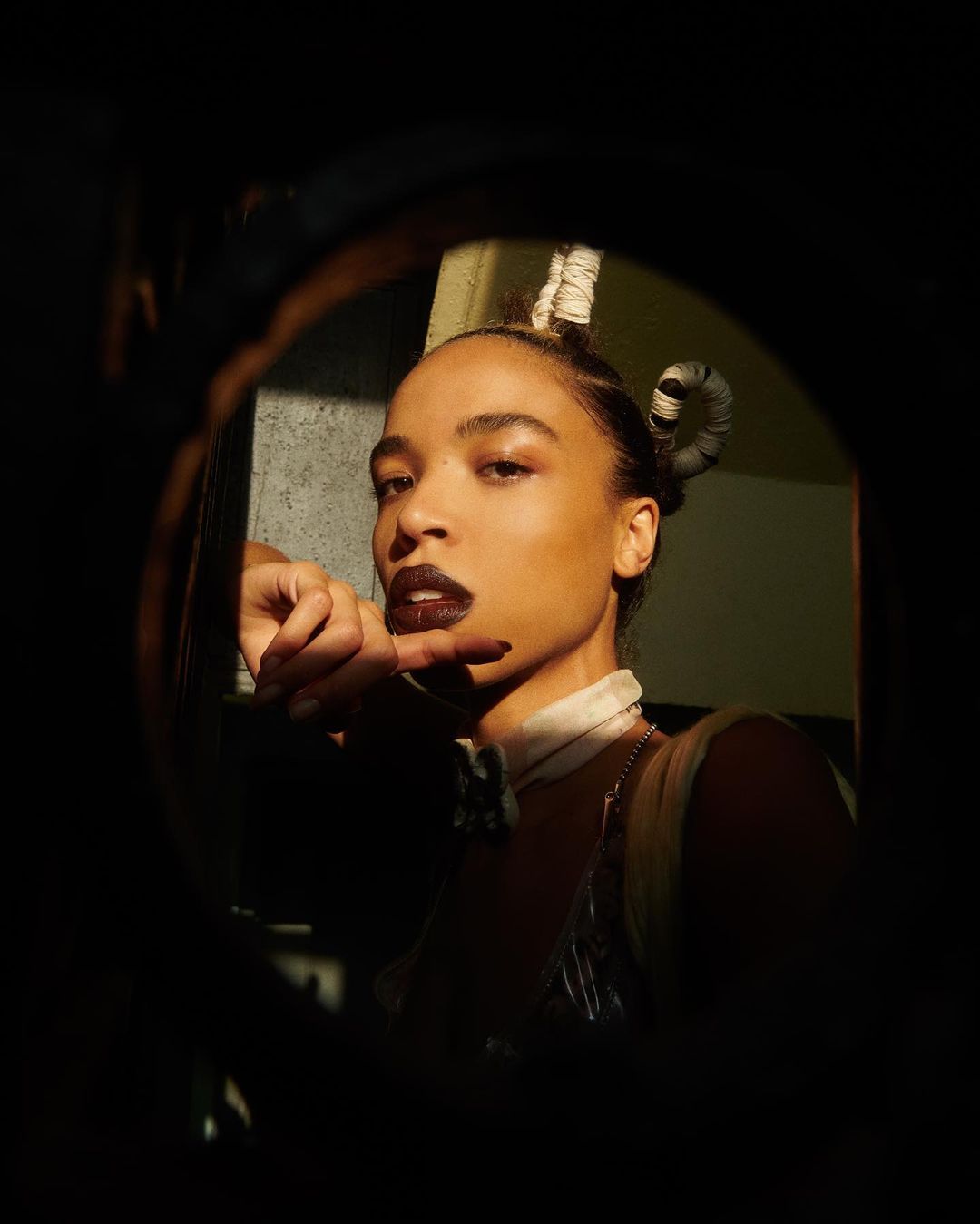 Portrait of an adult female with unique hair styling and dark lipstick, posing in a dark, enclosed space with dramatic lighting.