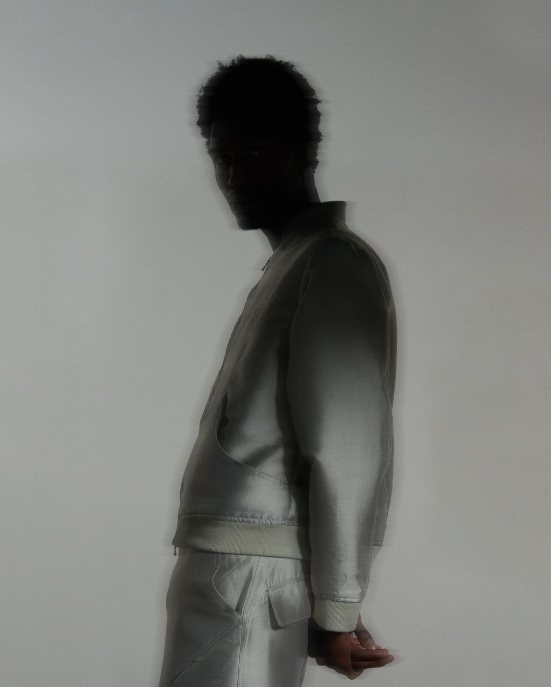 Side profile of an adult, silhouetted man wearing a pale jacket and pants against a muted background, soft focus, and studio lighting.