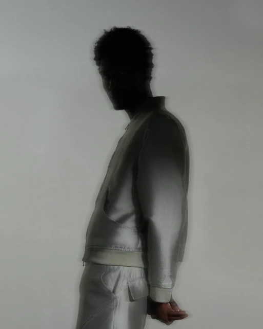 Side profile of an adult, silhouetted man wearing a pale jacket and pants against a muted background, soft focus, and studio lighting.