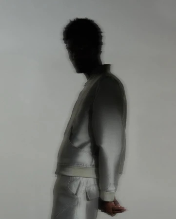 Side profile of an adult, silhouetted man wearing a pale jacket and pants against a muted background, soft focus, and studio lighting.