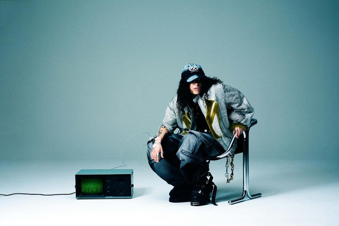 Young adult sits on metal chair next to vintage TV with denim cap and jacket in a minimalist studio setting for a fashion shoot.