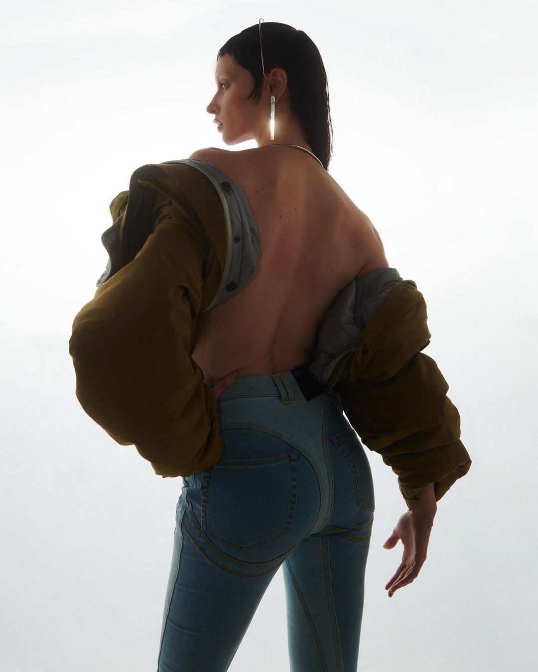 A woman with long dark hair poses from the back wearing jeans and a jacket with puffy sleeves against a bright white background.