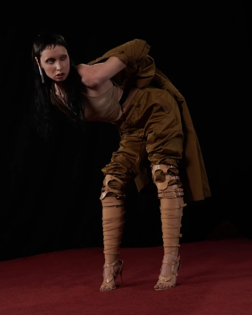 A woman poses in a unique outfit of wrap sandals, knee armor, loose pants and jacket in front of a dark background and red carpet.