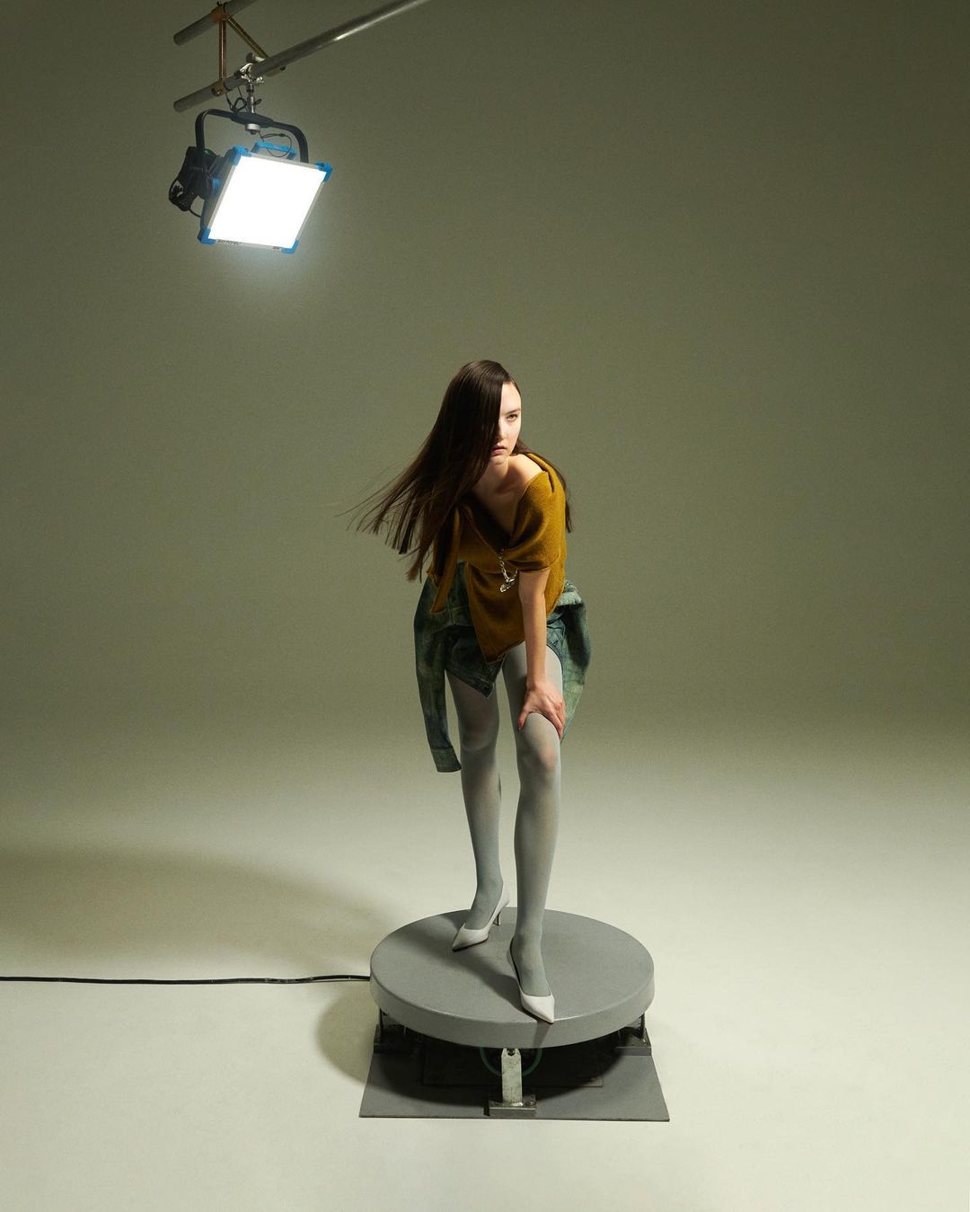 A female fashion model poses in a mustard-colored top, patterned skirt, and tights on a gray turntable in a studio setting with soft lighting.