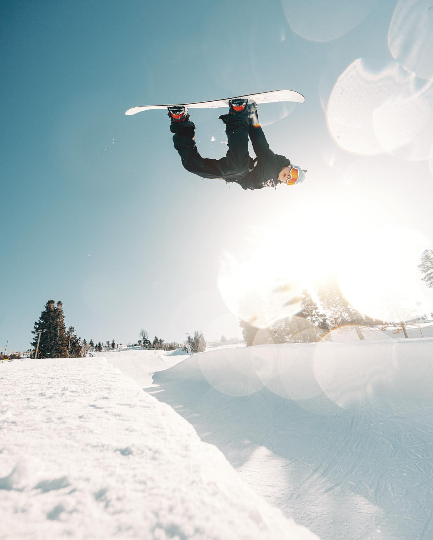 A snowboarder performs an inverted aerial trick high above a snowy slope on a bright, sunny day in an outdoor winter setting.
