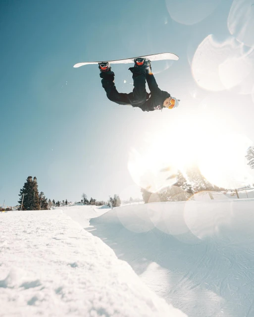 A snowboarder performs an inverted aerial trick high above a snowy slope on a bright, sunny day in an outdoor winter setting.